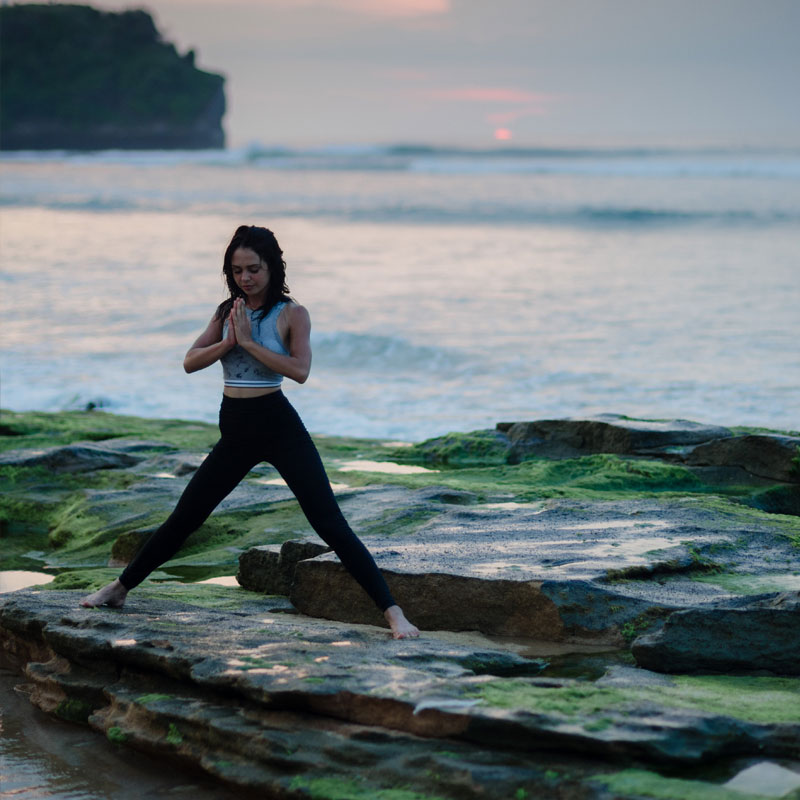 Woman doing yoga on rocks on the beach - representing the stressless benefits provided by a GoReady trip cancellation plan.