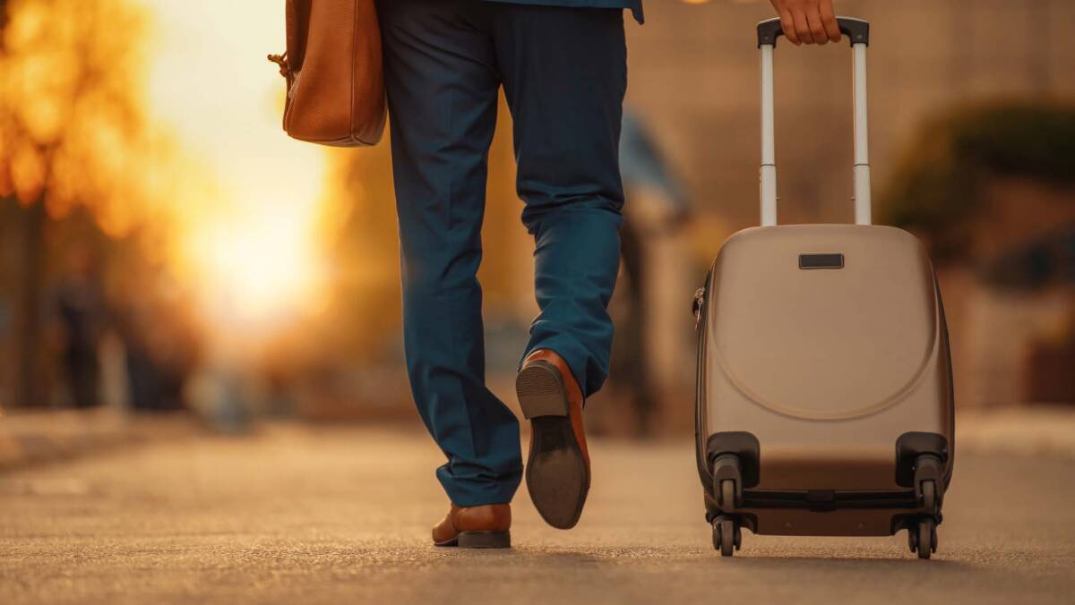 A man in a suit walking down the street holding luggage and backpack travel gear.